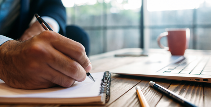 A male, wearing smart blue clothing, sat down, taking notes in a notepad, on a modern deck with a laptop and stationary on the desk.
