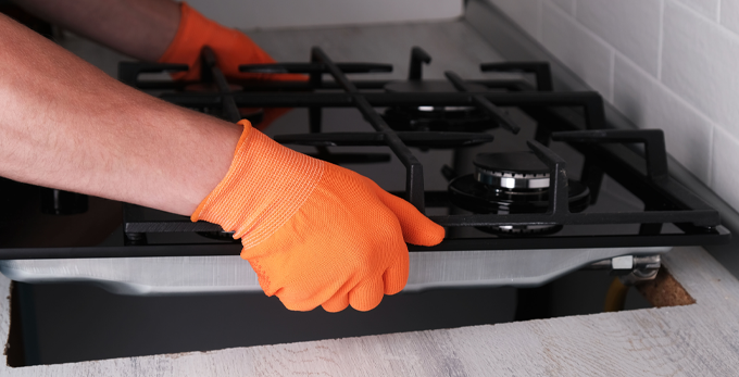 A plumber wearing protective orange gloves, installing a gas hob in a household kitchen.